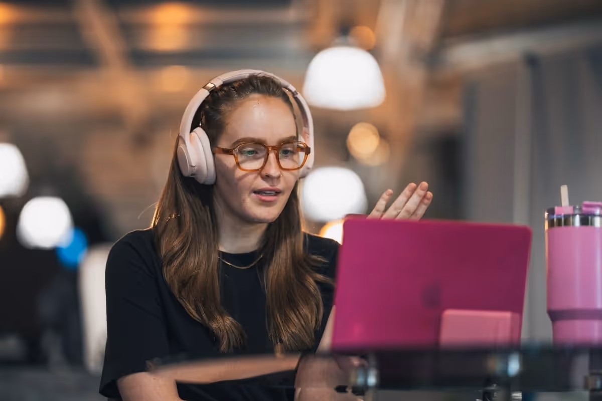 A woman in headphones and glasses talks to camera over her laptop in the office.