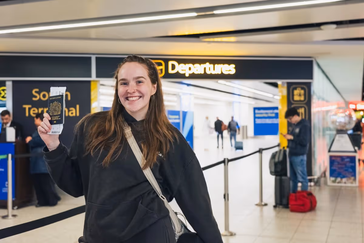 A smiling young woman holds her passport and boarding pass in the airport departures hall.