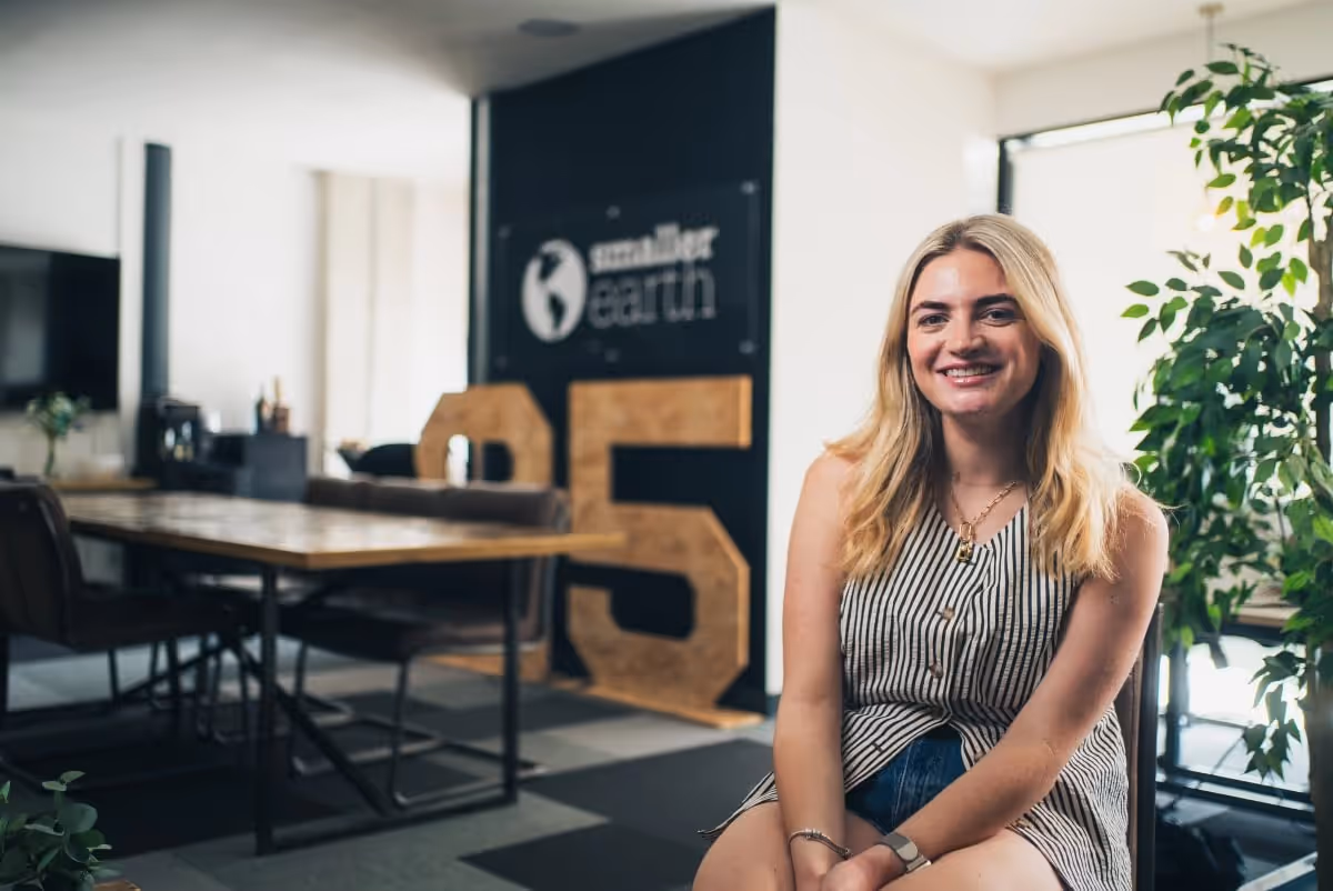 A woman sits for an interview at the Smaller Earth office, a large '25' anniversary sign behind her.