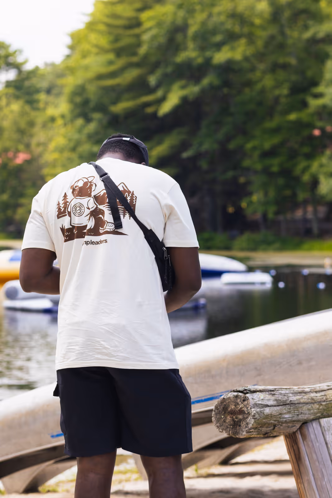 A crew member in a Camp Leaders t-shirt stands at the water's edge checking his camera bag, canoes and paddleboards moored behind him.