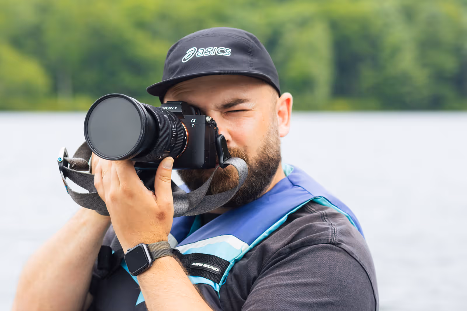 A bearded camera operator in a cap and buoyancy aid holds a mirrorless camera to his eye out on the lake.