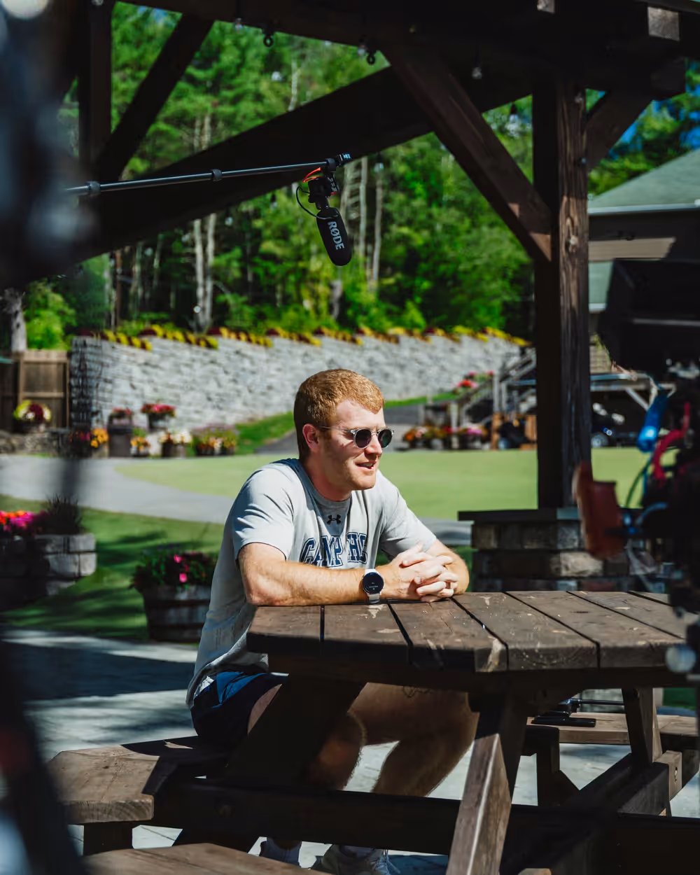 A camp director sits at a picnic table for a filmed interview, a boom microphone overhead and the camp lodge grounds behind him.