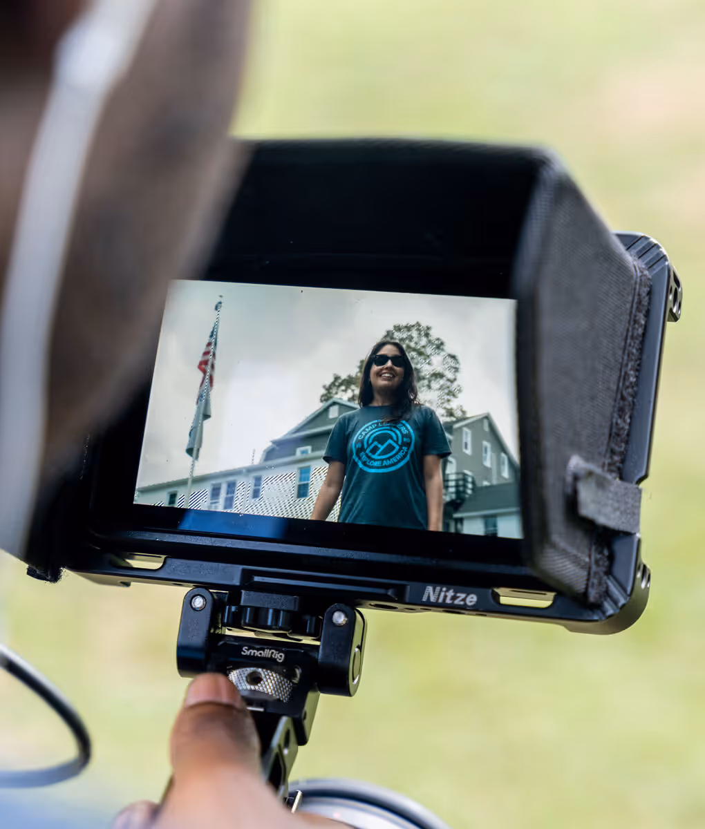 A camera monitor on set showing Immy smiling outside a camp lodge, the Stars and Stripes flying behind her