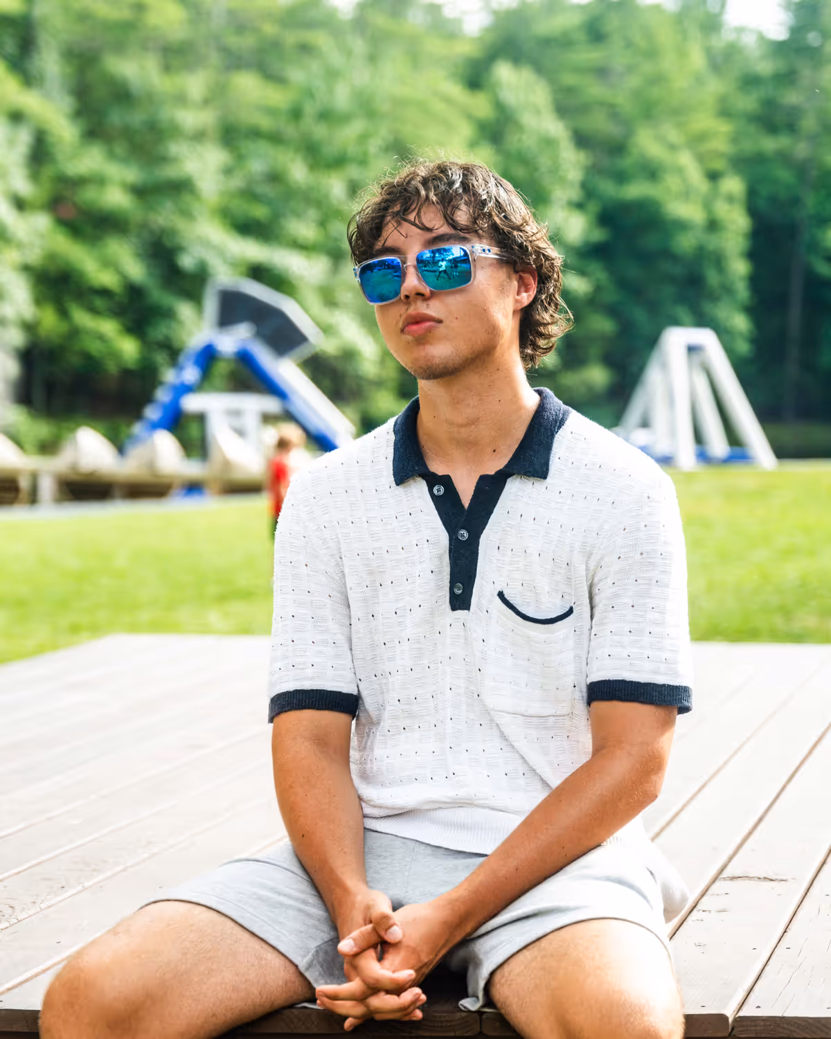 A young man in sunglasses sitting on a wooden deck at camp, the waterfront blurred behind him