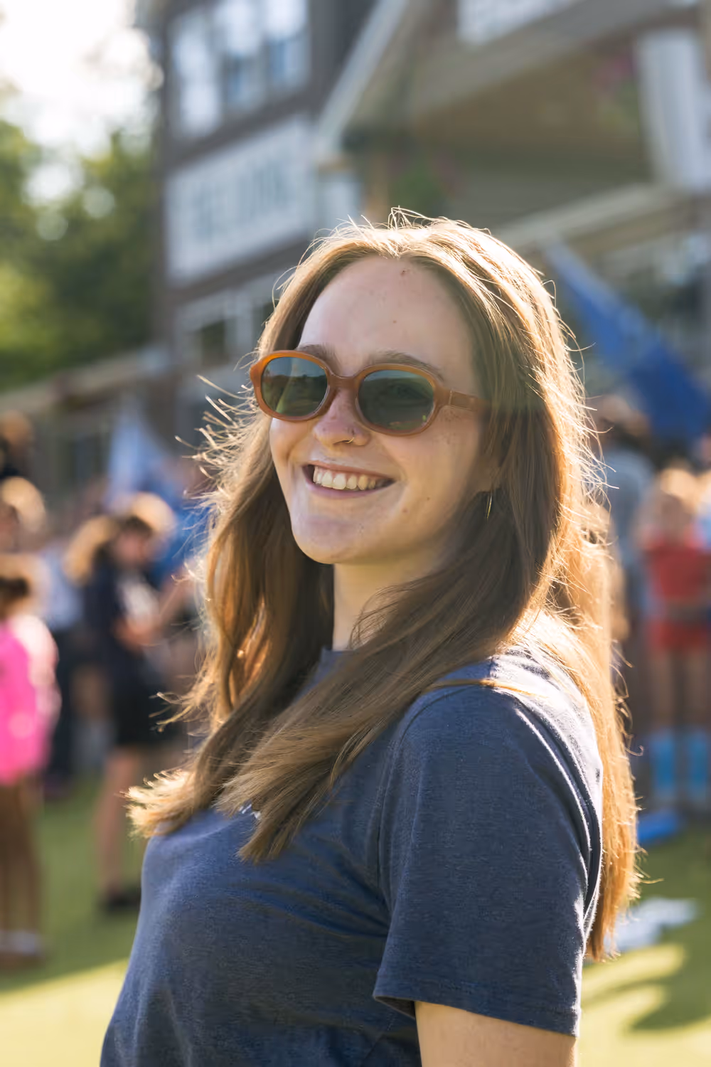 Immy, smiling in sunglasses with the late sun behind her, standing on the field as camp gathers in the background